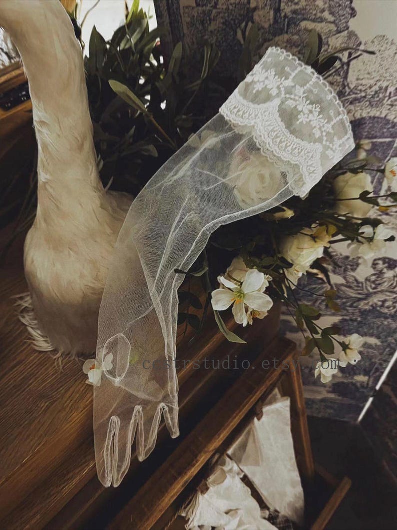 May include: A sheer white tulle glove featuring an intricate floral lace trim at the wrist. The glove is crafted from a delicate, semi-transparent mesh fabric and includes a circular cutout detail on the back of the hand, extending to the fingers.