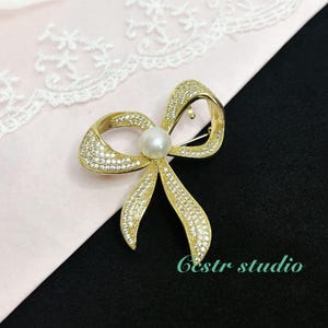 May include: A gold-tone bow brooch with clear rhinestones and a single pearl in the center. The brooch is on a pink background.