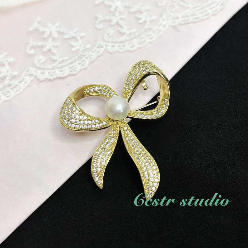 May include: A gold-tone bow brooch with clear rhinestones and a single pearl in the center. The brooch is on a pink background.