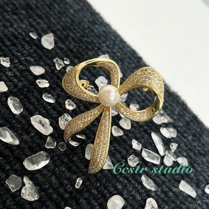 May include: A gold-tone bow brooch with clear rhinestones and a faux pearl in the center. The brooch is on a dark gray textured surface.