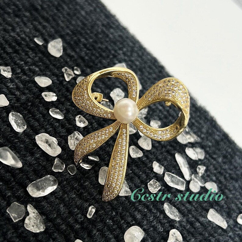 May include: A gold-tone bow brooch with clear rhinestones and a faux pearl in the center. The brooch is on a dark gray textured surface.