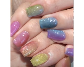 May include: Close-up of a hand with rainbow ombre glitter nail polish. The nails are short and square shaped.