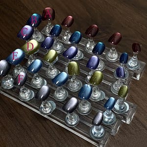 May include: A display of artificial nails in various colors, including blue, purple, green, and burgundy. Each nail is mounted on a clear stand, arranged in rows. The nails have a glossy finish, with some featuring glitter or a cat-eye effect. Letters A through F are visible.
