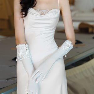 Könnte beinhalten: Ein weißes Satin-Trägerkleid mit Wasserfall-Ausschnitt und Spitzenbesatz sowie dünnen Spaghettiträgern. Das Set enthält lange weiße Satinhandschuhe mit Spitzenbündchen und drei dekorativen weißen Knöpfen am äußeren Handgelenk.