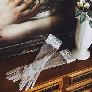 May include: A pair of sheer white tulle gloves featuring delicate floral lace trim at the cuffs. The gloves are translucent and extend to the mid-forearm, showcasing a fine mesh texture and intricate scalloped edges along the wrist openings.