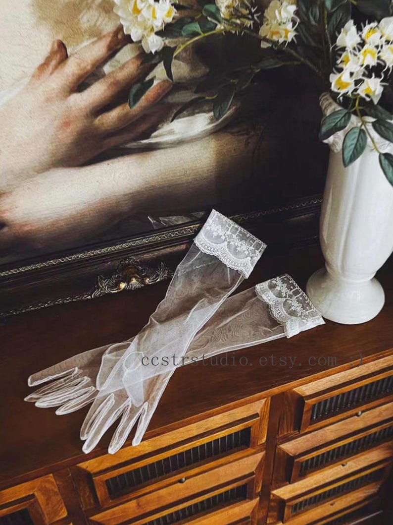 May include: A pair of sheer white tulle gloves featuring delicate floral lace trim at the cuffs. The gloves are translucent and extend to the mid-forearm, showcasing a fine mesh texture and intricate scalloped edges along the wrist openings.
