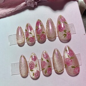 May include: Set of ten pink and gold false nails with a floral design. Each nail has a light pink base with a gold accent and a design of pink roses with green leaves.
