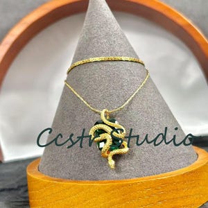 May include: A gold-colored necklace with a pendant featuring a green gemstone embraced by a snake design. The necklace is displayed on a gray cone-shaped stand. The image also shows a gold bracelet and the text "Ccstr studio".