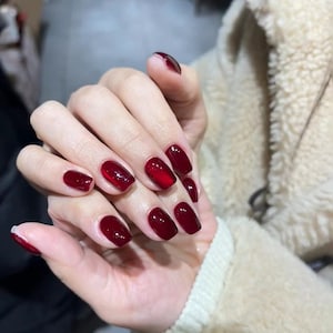 May include: A hand with polished nails. The nails are a deep, glossy red color, with a square shape. The manicure is well-manicured and the nails are all the same length. The background is blurred.