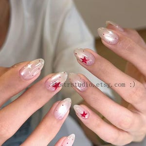 Könnte beinhalten: Nahaufnahme einer Hand mit oval geformtem, klarem Nagellack. Die Nägel haben ein perlmuttartiges Finish mit roten Stern- und Punktmustern. Der Text "strstudio.etsy.com :)" ist sichtbar.