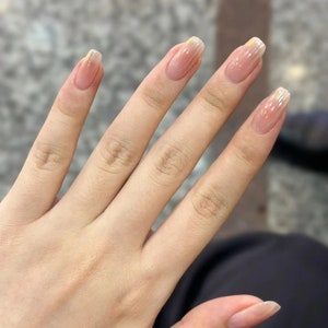 Könnte beinhalten: Nahaufnahme einer Hand mit fünf Nägeln, die mit einem hellrosa Nagellack mit einem dünnen goldenen Glitzerakzent an der Spitze lackiert sind.