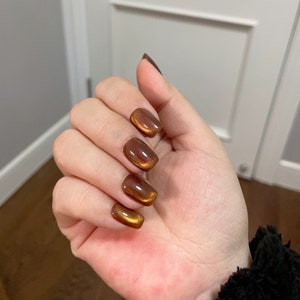 May include: Close-up of a hand with short, square-shaped nails painted with a shimmering, metallic brown nail polish.
