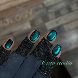 Peut inclure: Gros plan sur une main avec du vernis à ongles de couleur teal. Le vernis a un effet chatoyant, semblable à une galaxie. Les ongles sont de forme carrée. La main porte un gant sans doigts gris foncé. Le texte "Cestr studio" est visible.