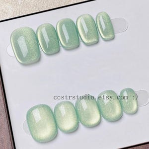Puede incluir: Un juego de diez uñas postizas en un tono verde menta claro brillante con un efecto ojo de gato resplandeciente. Las uñas están dispuestas en dos filas de cinco sobre una tarjeta blanca. El texto ccstrstudio.etsy.com :) está impreso en negro.