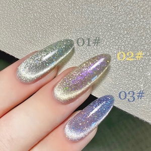 Puede incluir: Tres tonos diferentes de esmalte de uñas con purpurina. Los tonos son plateado, dorado y azul. El esmalte está en las uñas de una persona.