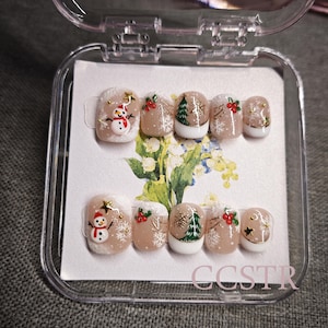 Könnte beinhalten: Ein Set mit zehn falschen Fingernägeln mit einem winterlichen Weihnachtsmotiv. Die Nägel sind mit Schneemännern, Weihnachtsbäumen, Schneeflocken und roten Beeren verziert. Die Nägel befinden sich in einem durchsichtigen Plastikbehälter mit weißem Hintergrund.