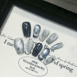 Könnte beinhalten: Ein Set mit zehn künstlichen Nägeln mit silbernem Glitzer-Finish und schwarzen Spitzen. Einige Nägel haben ein kleines silbernes Sternchen-Design. Die Nägel werden auf einer weißen Oberfläche mit einem kleinen schwarz-weißen Etikett mit der Aufschrift "@SiXi Wooden jewelry box NICE TO MEET YOU" präsentiert.