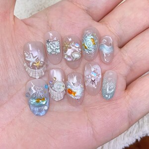 May include: A set of ten press-on nails featuring a light pink base with various 3D embellishments including miniature gold starfish, white seashells, iridescent pearls, silver foil accents, and small orange goldfish figures encased in clear resin.