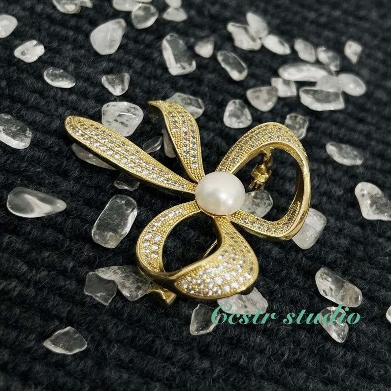 May include: A gold-tone bow brooch with a pearl in the center, surrounded by clear rhinestones. The brooch is on a black background with clear stones.
