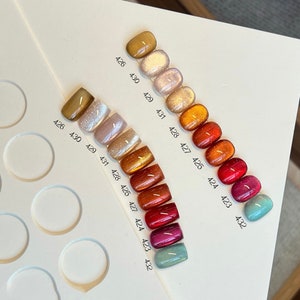 May include: A row of 12 small nail polish bottles with different shades of red, orange, yellow, and brown. The bottles are arranged in a line from left to right, with the darkest shade on the left and the lightest shade on the right. Each bottle has a number printed on it, starting with 426 and ending with 432.