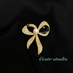 May include: A gold-toned bow brooch with clear rhinestones and a single white pearl in the center.