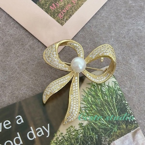 May include: A gold-toned brooch in the shape of a bow with clear rhinestones and a single white pearl in the center.