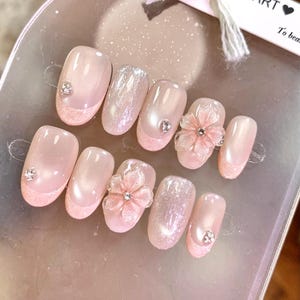 以下が含まれることがあります： ソフトピンクとパール仕上げの付け爪10本セット。グリッターのアクセント、小さな透明ラインストーン、立体的な花の装飾が施されています。ネイルはNAIL ARTと書かれた透明なプラスチックカードにディスプレイされています。