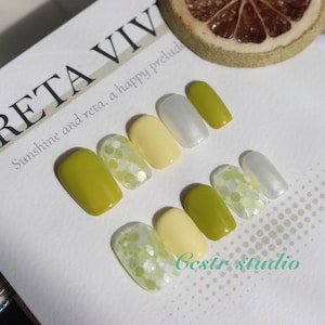 May include: Close-up of a set of ten press-on nails with a yellow and green color scheme. The nails have a glitter design and are arranged in a row on a white surface. The text "Sunshine and reta, a happy prelude" is visible in the background.