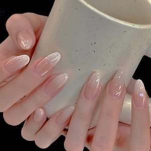 May include: Close-up of hands with long, oval-shaped, clear, light pink ombre nails. The nails have a subtle shimmer and are slightly translucent.