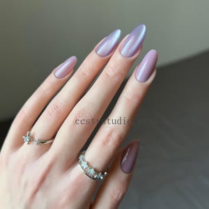 Könnte beinhalten: Eine Hand mit langen, oval geformten Nägeln, die in einem perlmuttartigen Lavendelton lackiert sind. Die Nägel haben ein glänzendes Finish. Zwei silberne Ringe mit Sternmotiven schmücken die Finger. Der Text "ccstystudio.etsy.com" ist enthalten.