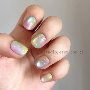 May include: Close-up of a hand with painted nails. The nails are a pastel rainbow gradient with iridescent shimmer and small star accents. The nail art features shades of pink, yellow, and blue.