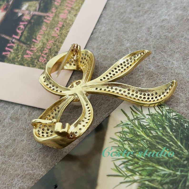 May include: A gold-toned bow brooch with a sparkling, clear stone in the center. The bow is made of metal with a textured surface and has a pin on the back for attaching to clothing.