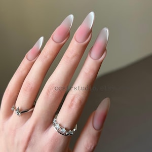 以下が含まれることがあります： アーモンド型の人工爪で、ソフトピンクから白のグラデーションデザイン。爪は長く光沢があり、滑らかな仕上がりです。星と三日月をモチーフにした2つのシルバーリングも写っています。
