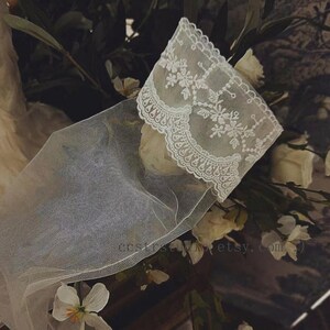 May include: A sheer white tulle glove featuring an intricate floral lace cuff with scalloped edges. The delicate fabric is semi-transparent, showcasing detailed embroidery patterns along the wrist area. The text costumestudio.etsy.com is visible.