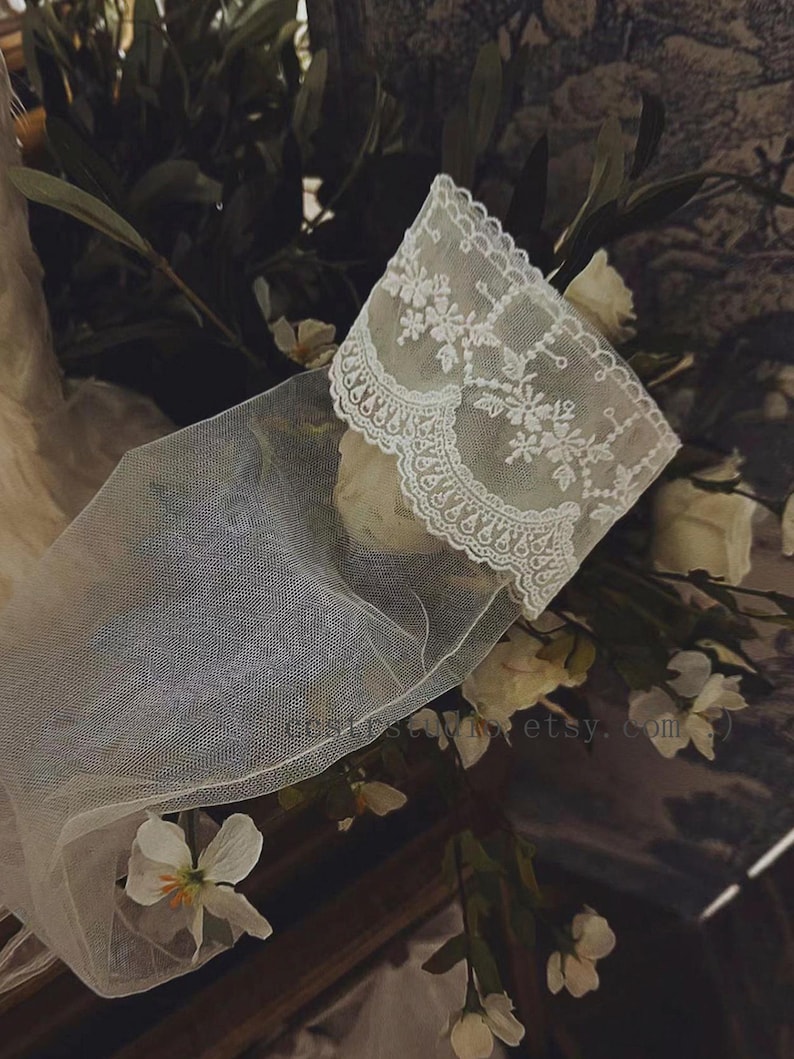 May include: A sheer white tulle glove featuring an intricate floral lace cuff with scalloped edges. The delicate fabric is semi-transparent, showcasing detailed embroidery patterns along the wrist area. The text costumestudio.etsy.com is visible.