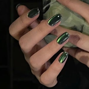 May include: Close-up of a hand with polished nails. The nails are a glossy, dark green color with a cat-eye effect, creating a shimmering, reflective appearance. The nails are oval-shaped and well-manicured, with a smooth, even surface.