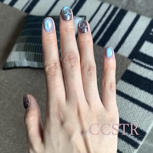 May include: A hand with five fingers, each with a different nail polish color. The nails are decorated with small, silver stars. The hand is resting on a striped surface.