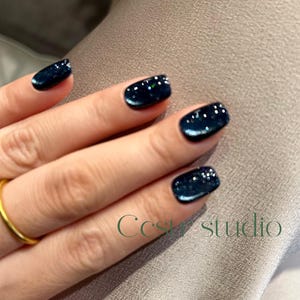 Pode incluir: Close-up de uma mão com verniz de unhas azul escuro com glitter. As unhas são curtas e quadradas. Um anel de ouro está no dedo anelar. O texto "Ccs studio" está em verde.