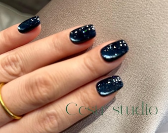 Könnte beinhalten: Nahaufnahme einer Hand mit dunkelblauem Nagellack mit Glitzer. Die Nägel sind kurz und quadratisch geformt. Ein goldener Ring befindet sich am Ringfinger. Der Text "Ccs studio" ist grün.