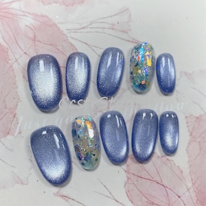 May include: A set of ten artificial nails in a light blue hue. Some nails have a silver shimmer effect, while others feature iridescent glitter. The nails are oval-shaped and arranged on a light pink surface.