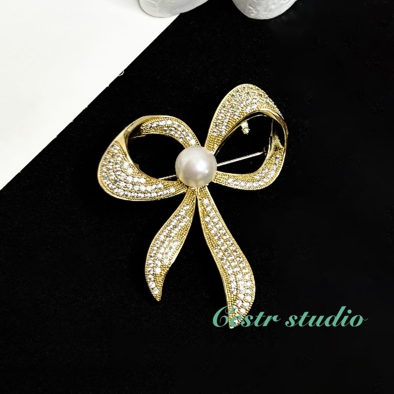 May include: A gold-toned bow brooch with a pearl in the center, adorned with sparkling rhinestones.