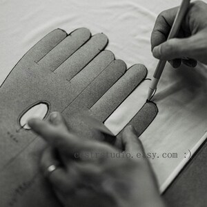 May include: A gray cardboard glove pattern template with finger outlines and a small oval cutout. A pen is used to trace the outline onto white fabric. The text cstrstudio.etsy.com is printed on the template surface in a black sans-serif font.