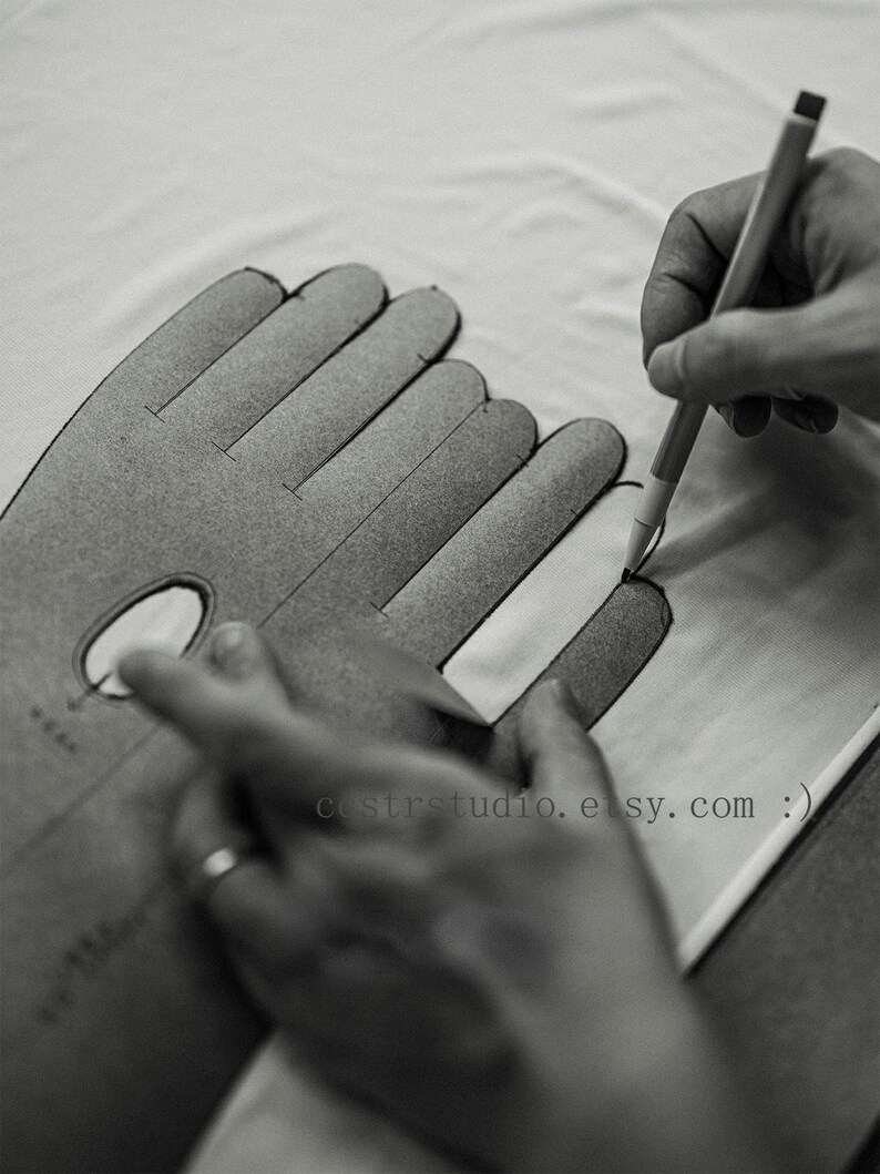 May include: A gray cardboard glove pattern template with finger outlines and a small oval cutout. A pen is used to trace the outline onto white fabric. The text cstrstudio.etsy.com is printed on the template surface in a black sans-serif font.