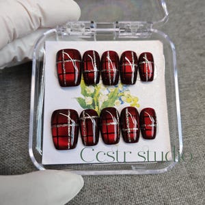 Könnte beinhalten: Ein Set künstlicher Fingernägel in einer transparenten Kunststoffbox. Die Nägel sind tiefrot mit einem Karomuster in Schwarz und Silber. Der Text "Ccstr studio" ist auf der Karte im Inneren der Box zu sehen.