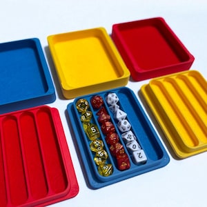 Pode incluir: Uma coleção de bandejas coloridas para dados em azul, amarelo e vermelho. Uma bandeja azul contém uma variedade de dados, incluindo dados transparentes, vermelhos e brancos. As bandejas são projetadas para organizar e guardar dados.