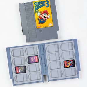 Puede incluir: Un cartucho gris de Nintendo con la etiqueta "Super Mario Bros. 3" se encuentra sobre un soporte gris para cartuchos de juego. El soporte está abierto, revelando varios cartuchos de juego. Algunos están insertados.