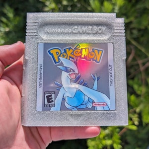 May include: A silver Game Boy cartridge for the game Pokémon. The label features the Pokémon logo and a blue and white illustration of a creature. The cartridge has the text "THIS SIDE OUT".