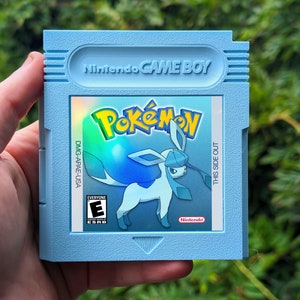 以下が含まれることがあります： ポケモンゲームの、水色のニンテンドーゲームボーイカートリッジ。ラベルには、ポケモンのロゴと、青と白のグレイシアのキャラクターが表示されています。カートリッジには「Nintendo GAME BOY」と「THIS SIDE OUT」の文字があります。