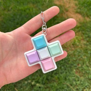 Peut inclure: Un porte-clés en forme de croix aux couleurs pastel, avec une chaîne argentée. Le porte-clés présente une bordure blanche et des carrés dans les tons de bleu, vert, rose et violet. L'arrière-plan est de l'herbe verte floue.