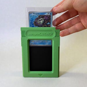 May include: A green "CARD BOY" card holder, resembling a handheld game console, with a Pokemon card inside. The text "wookieeprint CARD BOY" is printed on the holder. A Pokemon card is held in a clear plastic sleeve. The holder is green.